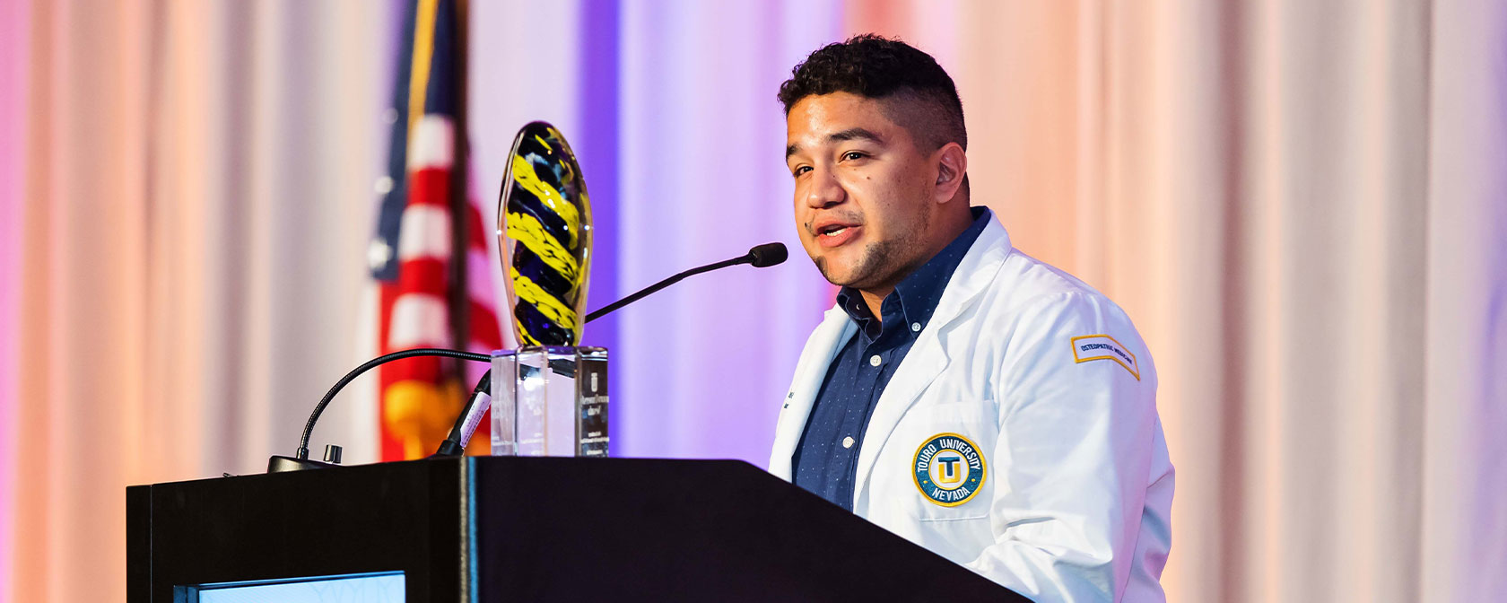 Medical student, Jose Parra, in his white coat, speaks at the podium during the Gala 2022 event.