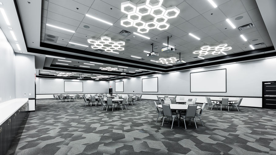 A large conference room with round tables surrounded by chairs, projector screens along the walls, and a long counter on the left side wall.