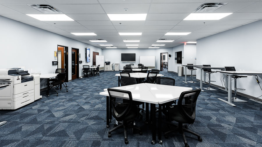 A large room with modular desks surrounding by office chairs in the center of the room. Tall tables with chairs line the right wall, and a copier and more desk line the left. Along the back wall is a TV monitor and two white boards.