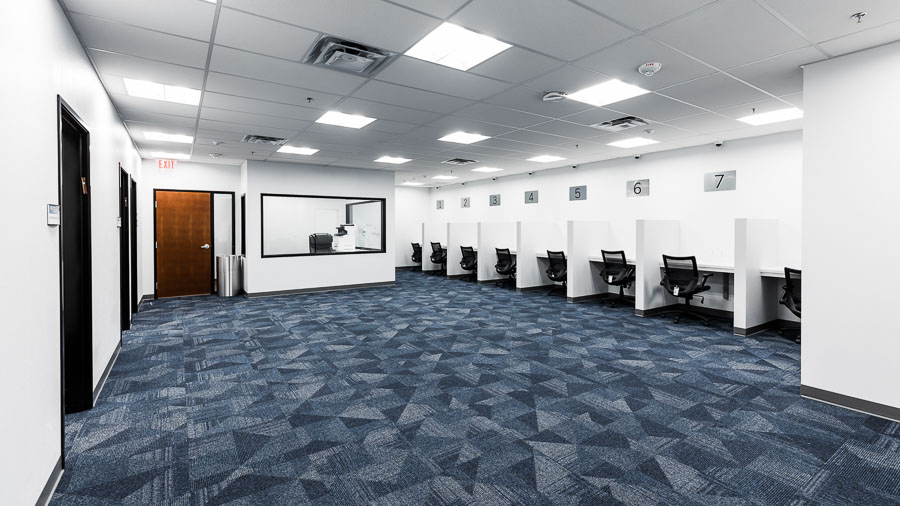 A large room with numbered cubicles along the right wall. Each cubicle contains and empty desk and an office chair. Against the back wall is a door to exit and a small observation booth with a window.