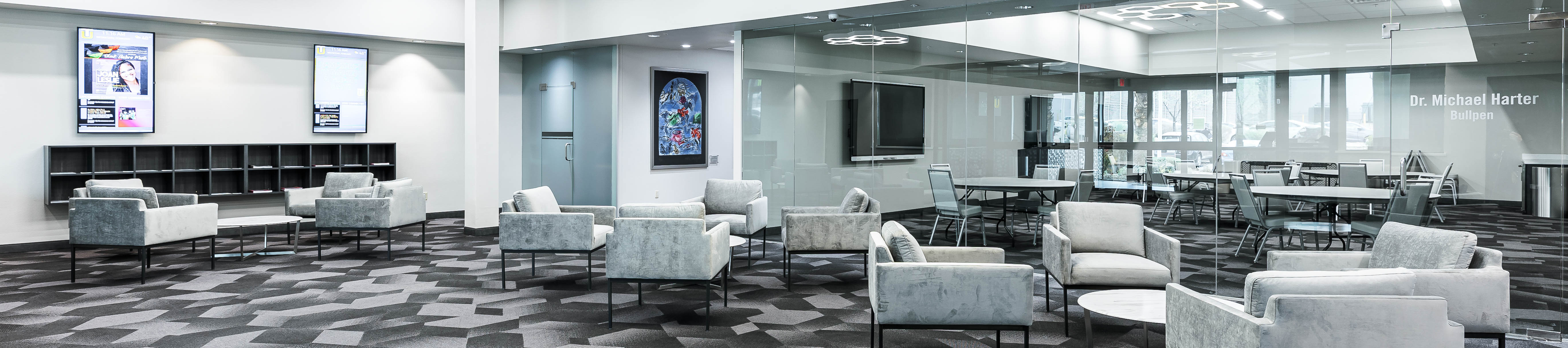 An empty front lobby with low circular tables surrounded by chairs. To the left is a wall of cubbies and two vertical monitors displaying university information. The back wall is made of glass, and shows a secondary conference room with tables, chairs, and TV monitors on the walls.