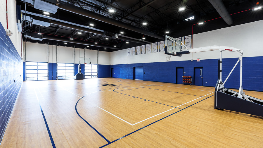 An indoor court with windows containing two basketball hoops on either end, and a rack of basketballs along the back wall.