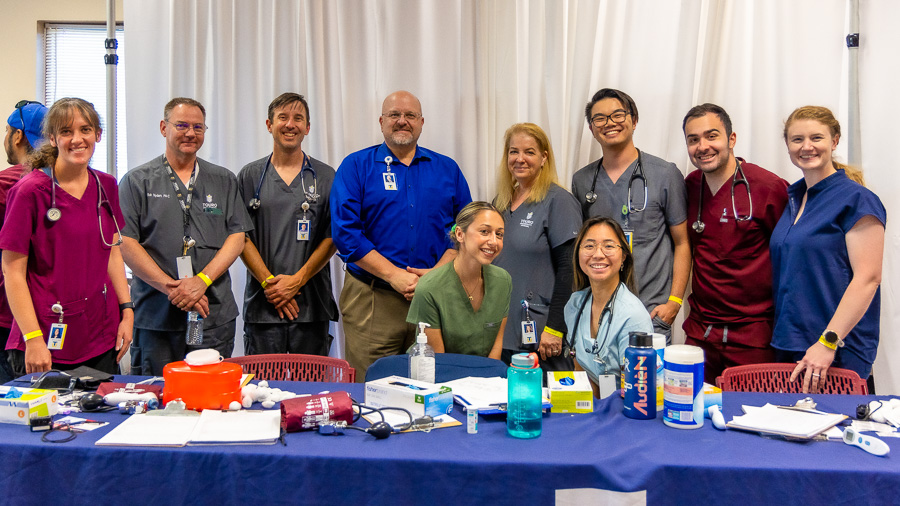 President Dr. Priest stands with faculty and staff, dressed in scrubs, behind a table covered in medical supplies.