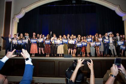 DO students on stage hold signs showing their residency placements as the audience photographs them during the Match Day event.