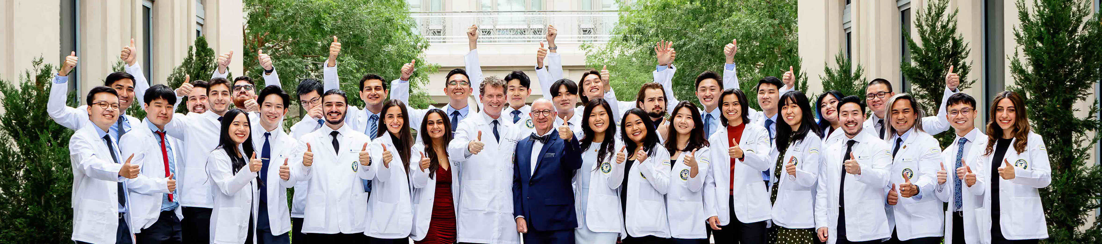Dr. Gilliar and Dr. Tompkins in the center of a group of Osteopathic Medicine students in an outdoor courtyard. The students wear their white coats gesture a thumbs up.
