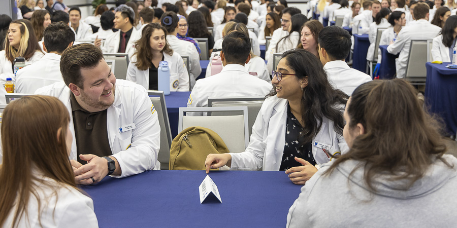 Students in white coats sit at tables and talk during an Interprofessional Education event.
