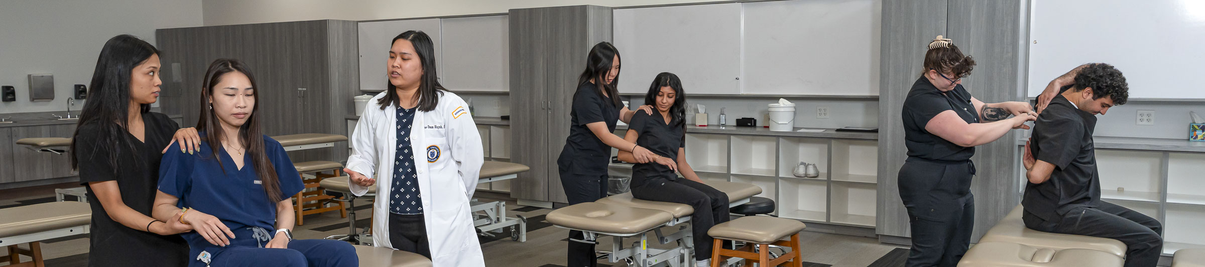 Students in scrubs practice examining each other’s shoulders and arms while a faculty member guides a pair of them.