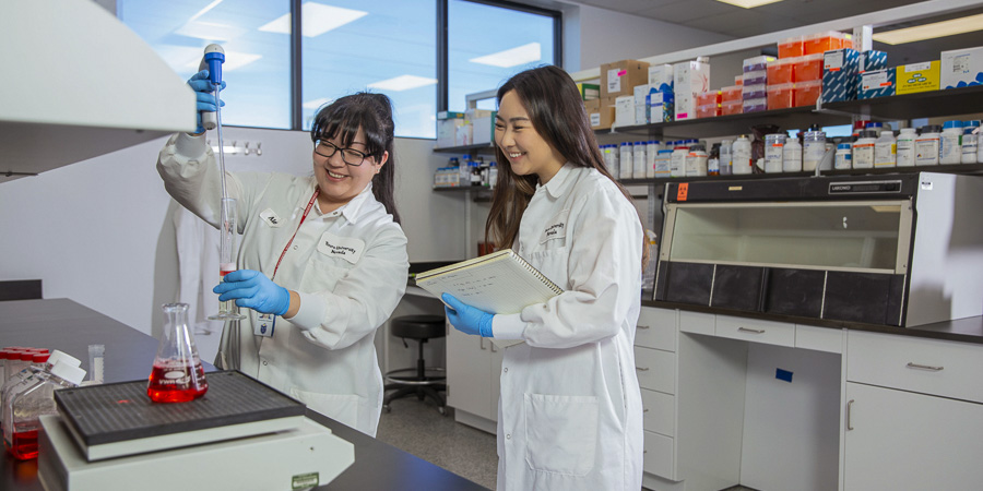 Two students in white lab coats and gloves work in a laboratory. One uses a pipette to transfer liquid into a graduated cylinder while the other holds a notebook.