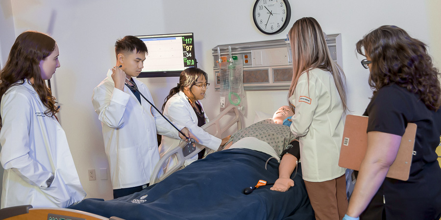 An instructor and students in white coats gather around a patient simulation manikin in a clinical skills lab while one student uses a stethoscope.