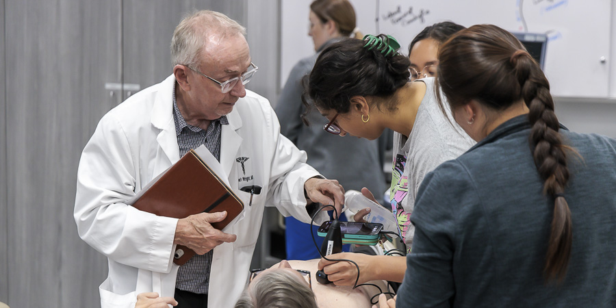 An instructor in a white coat guides students as they use a handheld ultrasound device on a patient in a clinical skills lab.