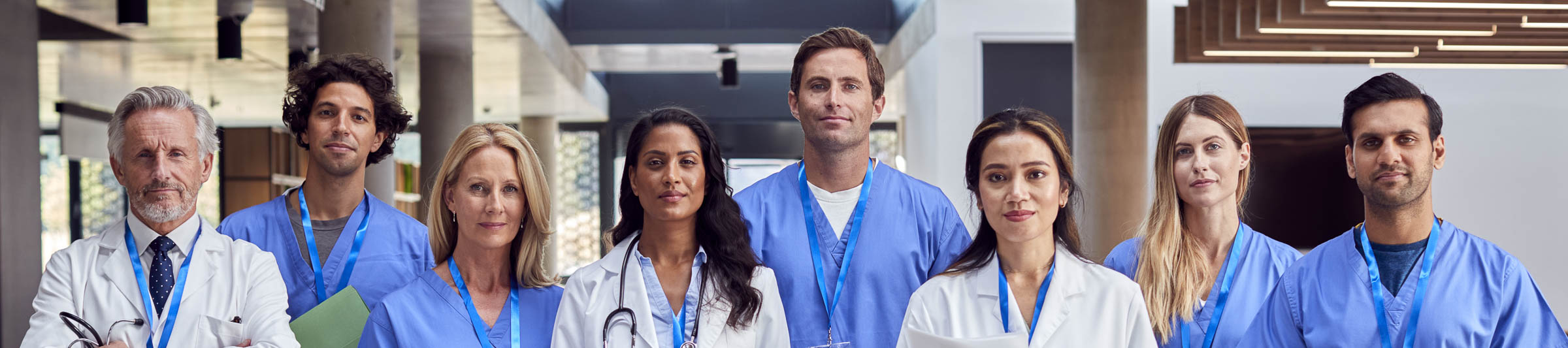A group of healthcare professionals in scrubs and white coats standing in a lobby, wearing badge lanyards and holding stethoscopes and paperwork.