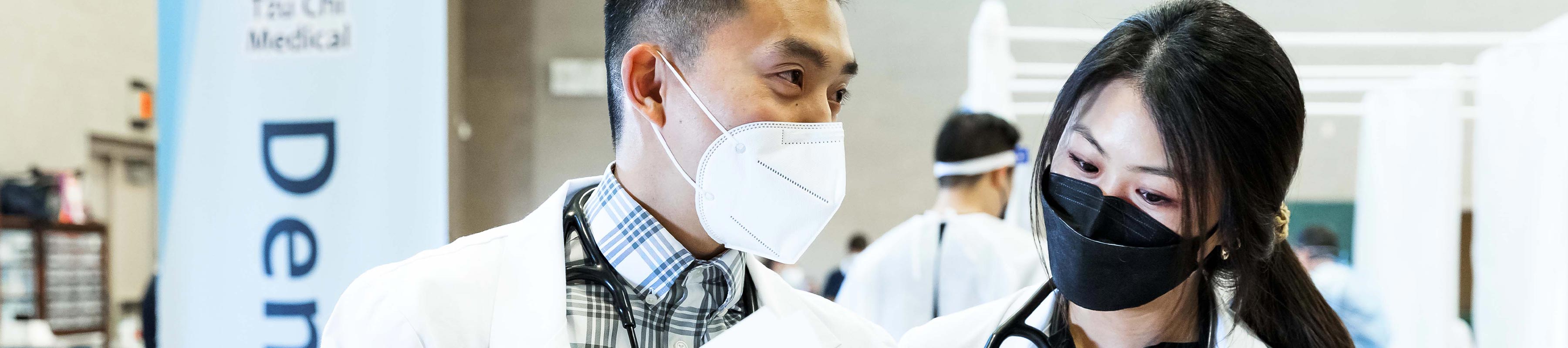 Two masked students conversing in their white coats at a community healthcare event.
