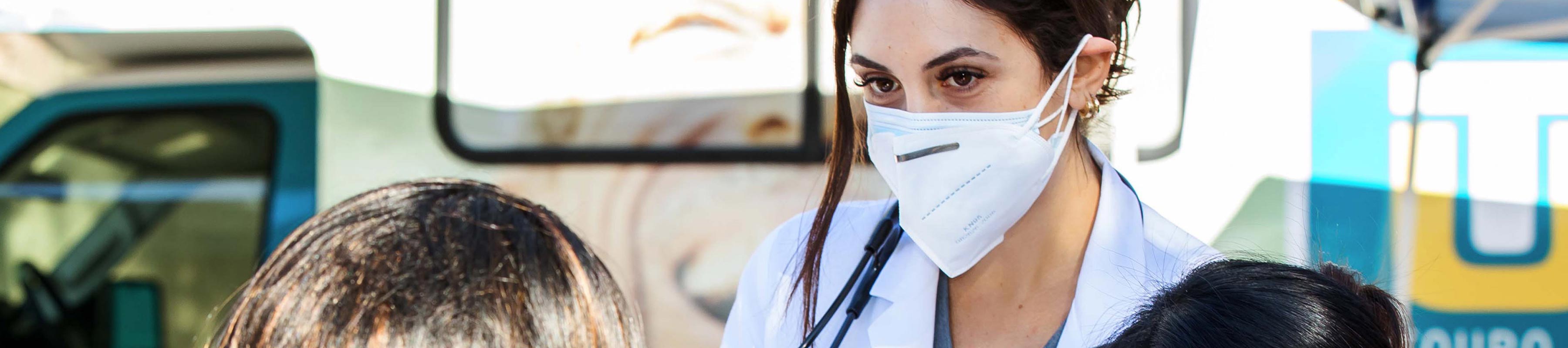 Two masked students in white coats talking with a patient