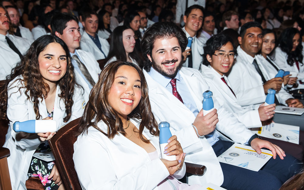 A group of medical students wearing white coats hold up their personal VAVE ultrasound devices from their seats during the White Coat Ceremony