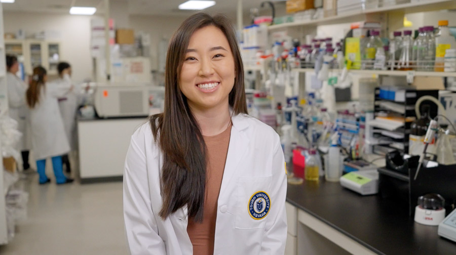 A woman in a white lab coat smile at the camera from the middle of a research lab. In the background, long counters are filled with lab equipment.