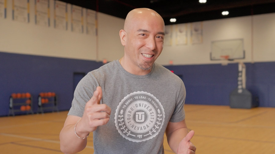 A man smiles and points to the camera from the center of an indoor court. In the background, to the left, are racks of basketballs, and to the right is a basketball hoop.