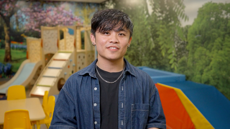 A man smiles at the camera from the middle of a large room filled with children's play equipment, including a small wooden jungle gym and child size table. The back wall of the room is covered in a large mural of trees and flowers.