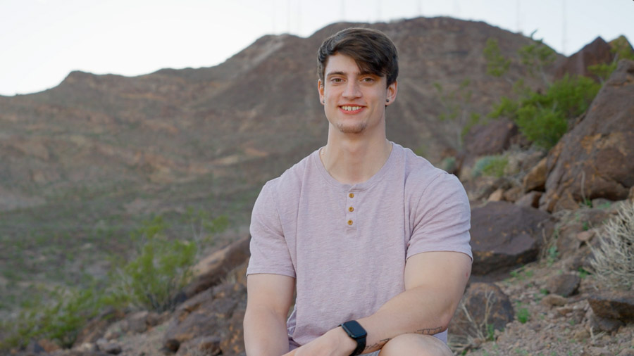 A man sits on a rock at the base of a mountain. In the background is a large mountain and desert landscape.