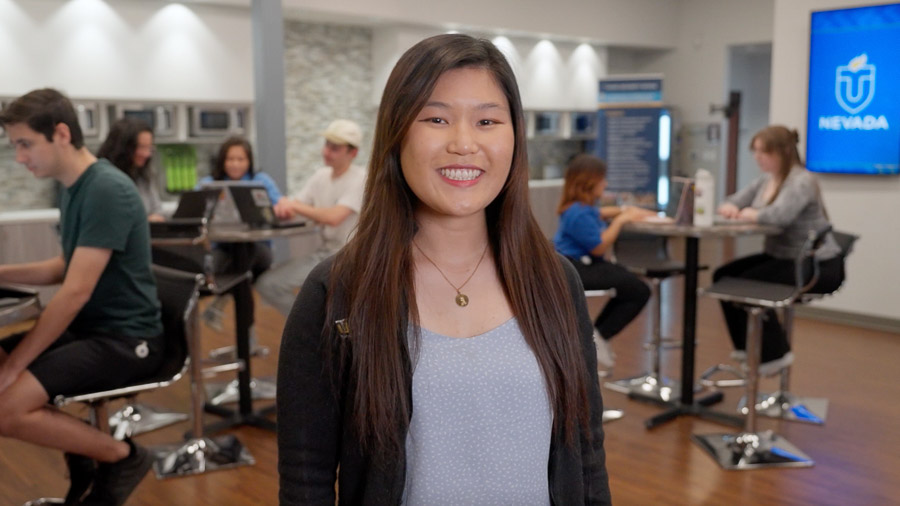 A woman smiles at the camera from the center of a student lounge. Students sit around high tables on either side of her studying. In the background is a long counter with coffee machines and microwaves and a TV monitor showing the Touro Nevada logo.