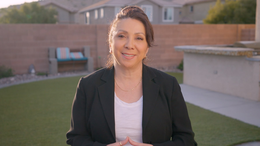 A woman smiles at the camera from a backyard with artificial turf and an outdoor kitchen. Beyond the block wall surrounding the yard are houses and mountains.