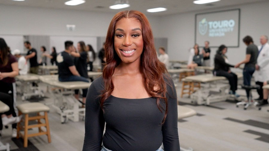 A woman smiles at the camera from the edge of a class room filled with exam tables. In the background, groups of medical students perform practice examinations on one another.