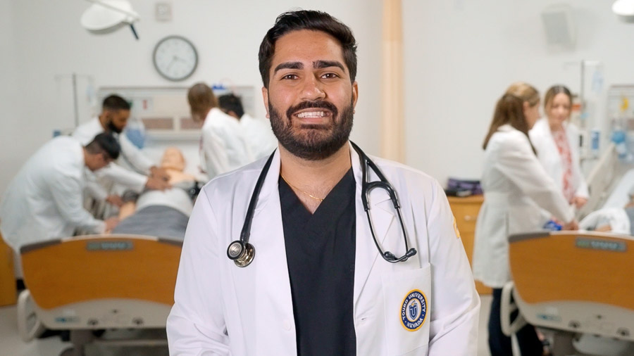 A man wearing a white coat and stethoscope stands in a simulated medical facility, smiles at the camera. In the background, medical students work on training manikins in hospital beds.