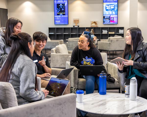 Five students holding lap tops and tablets engage in conversation around a table in a student common area.