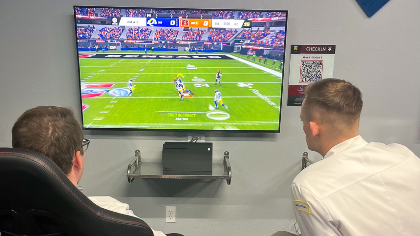 A students sit in front of a large screen monitor playing an Xbox X console game. Another student sits in a chair watching the game.