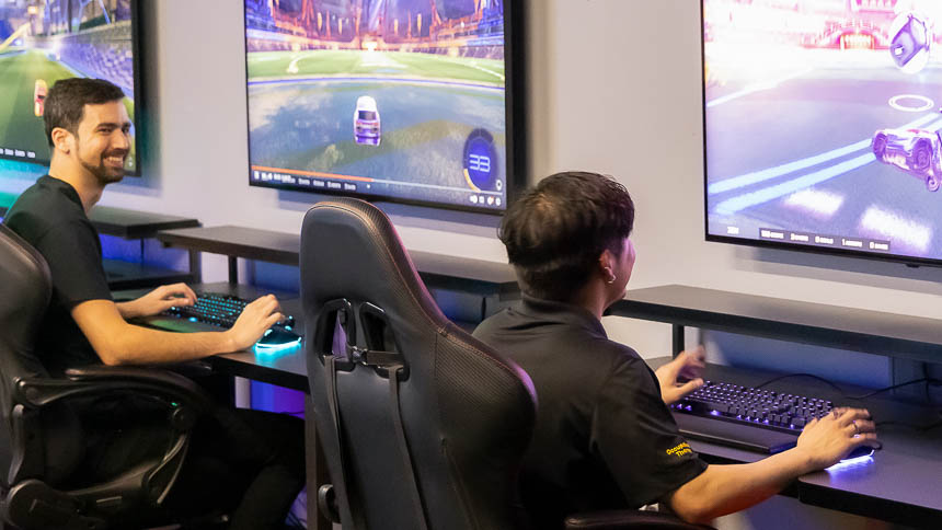 Two students sitting next to each other at desks playing a car racing PC game on large screens.