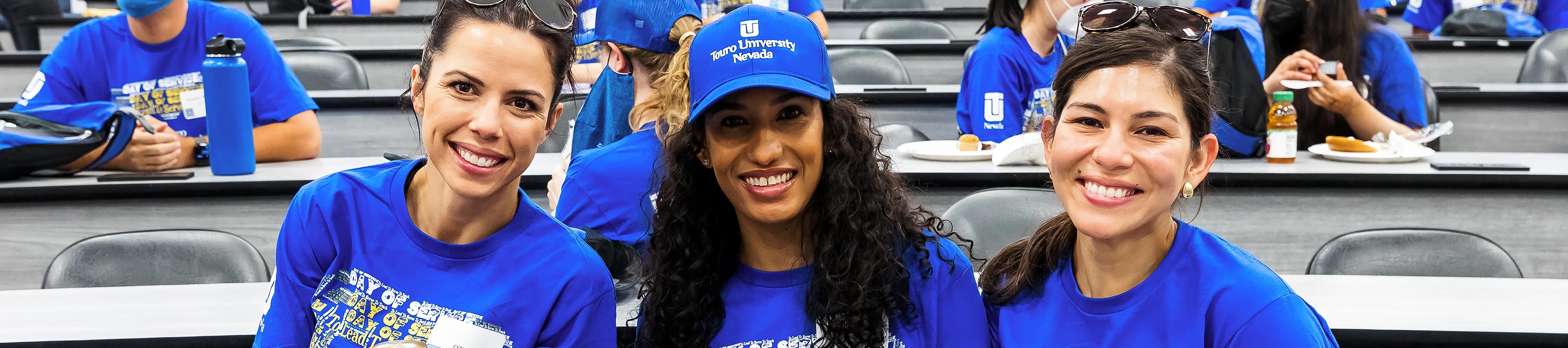 Three students in Touro hats and shirts smiling in a lecture hall.