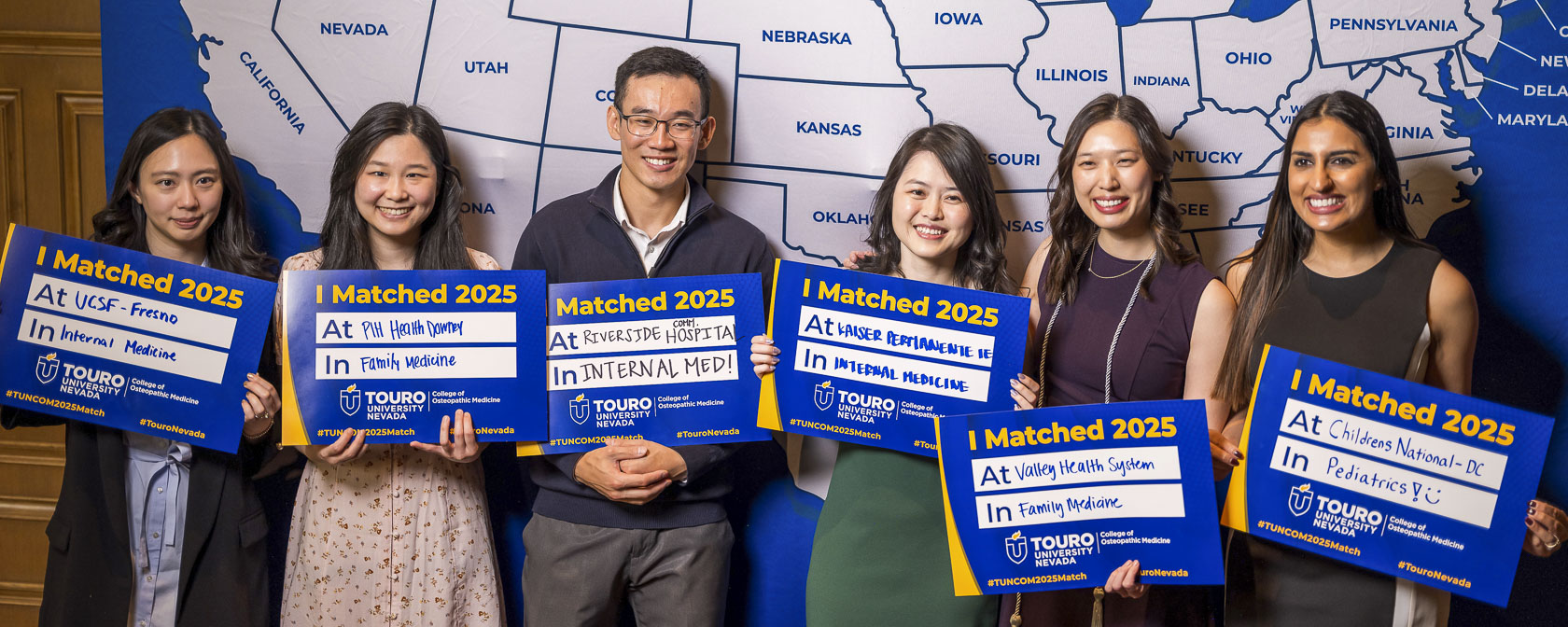 DO students pose for a group photo in front of a large map of the United States, each holding a sign displaying their residency location and specialty