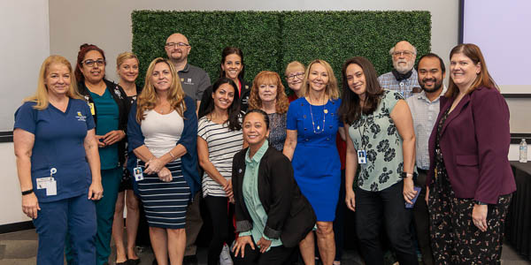 A group of 5 year employees gather for a group photo in front of a greenery wall.