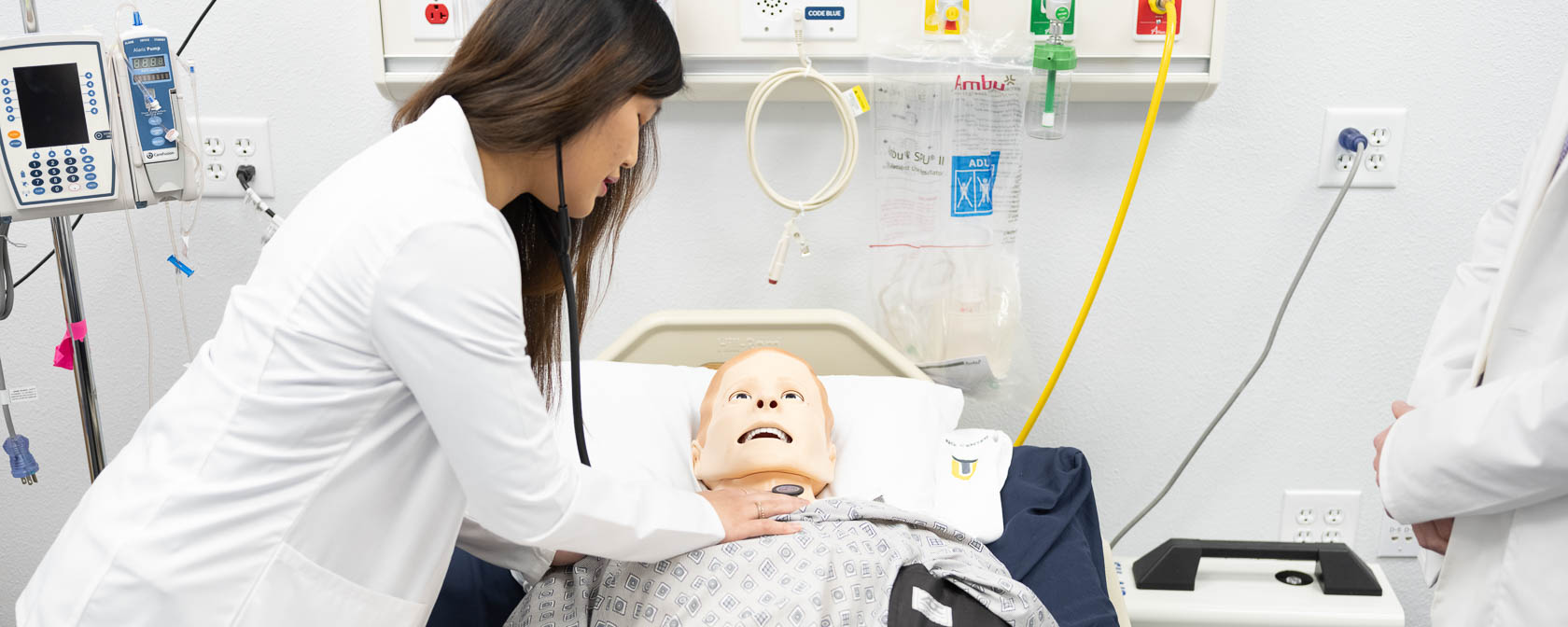 A College of Osteopathic Medicine student uses a stethoscope to listen to the chest of a manikin in a simulation center hospital bed.