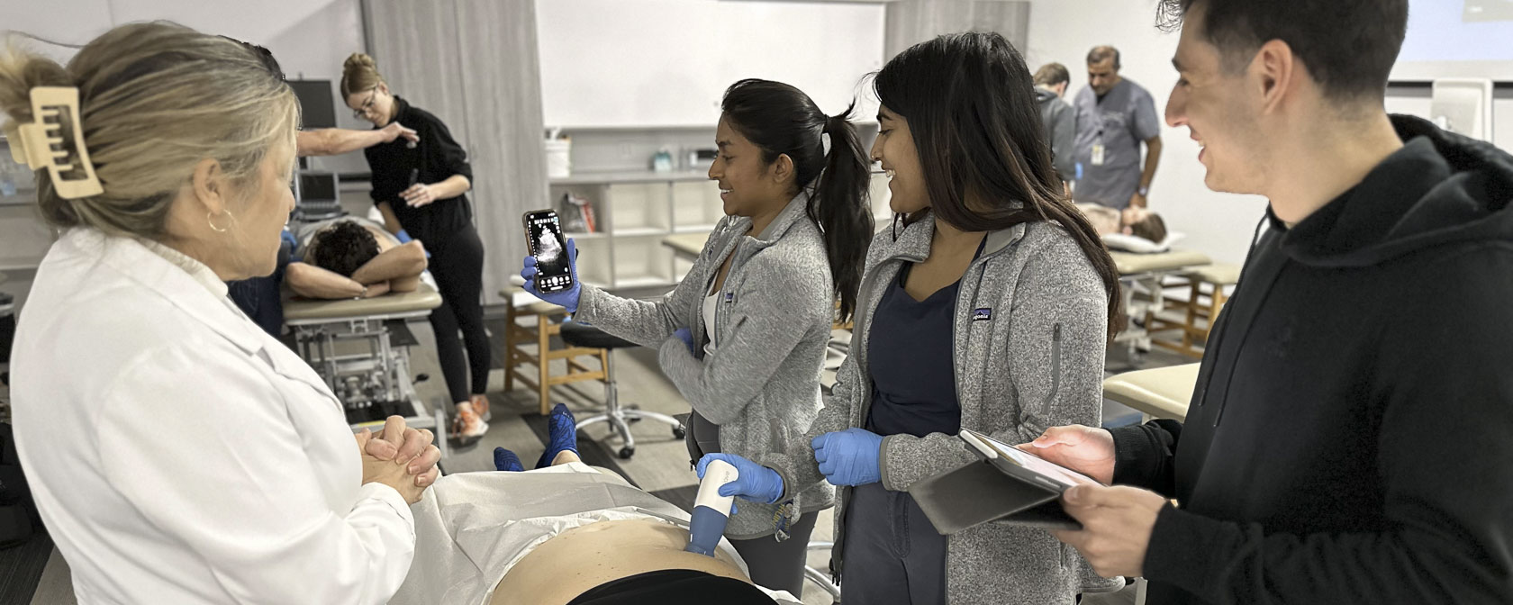 Students perform a point-of-care ultrasound on a patients abdomen while observing the results on a mobile device.