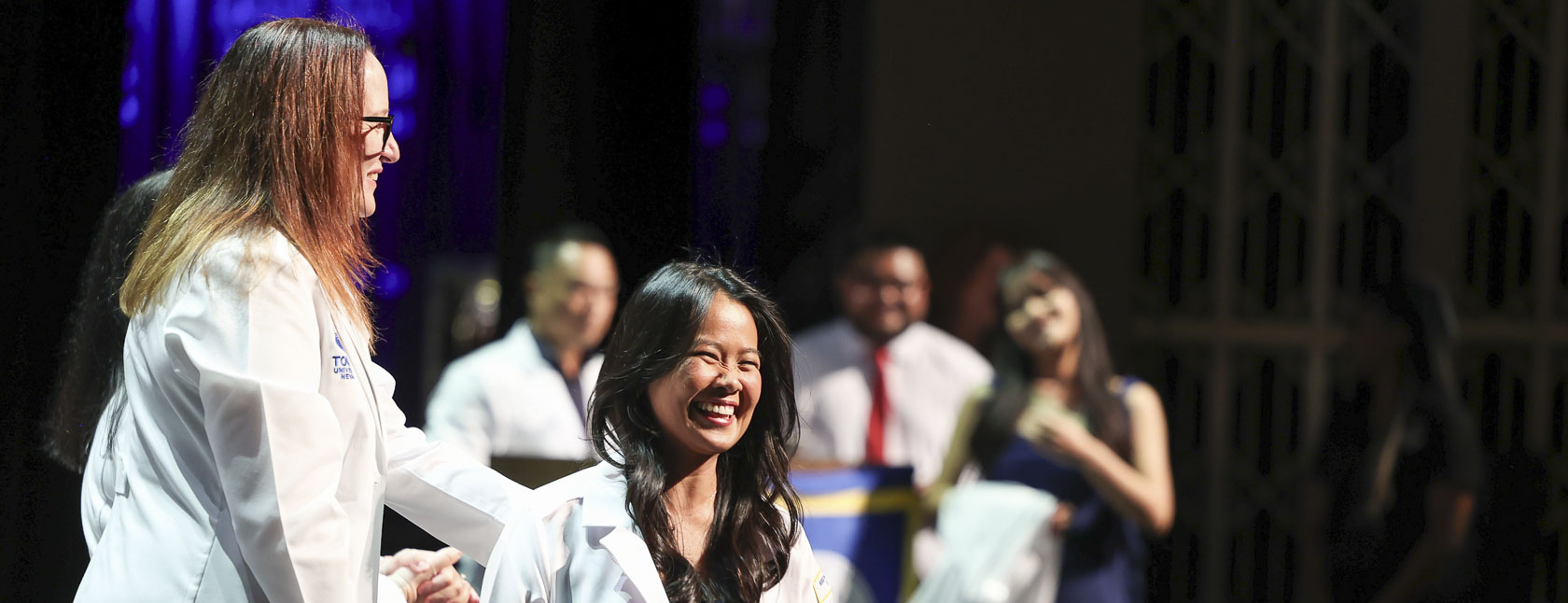 A Touro Nevada faculty member helps a student into her white coat on stage during the White Coat Ceremony.